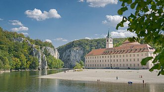 Flussufer mit Kiesstrand, altes Gebäude mit Turm und bewaldete Felsen unter blauem Himmel mit Wolken
